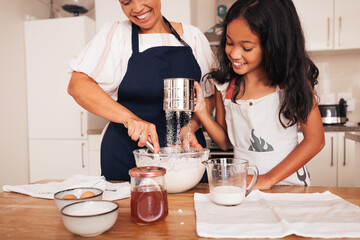 Senior woman and granddaughter sifting flour at a table in kitchen. Girl and her grandma making...