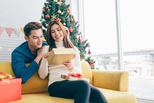 A happy Asian man surprises his girlfriend with Christmas presents at home with a Christmas tree in the background. image with copy space