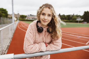 Young girl leaning on railing with headphones in front of sports field