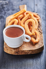 Chocolate en una taza de color blanco sobre una superficie rústica y rodeada de churros y un fondo negro