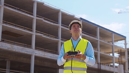 Drone operator holding remote controller. Professional builder in helmet and vest standing in front of construction site. Office building and crane background. Business, real estate and investment.