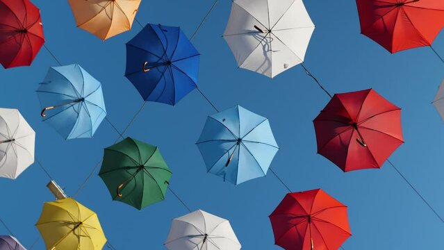 Multicolored umbrellas hang against the blue sky and rotate. The smooth departure of the camera. Urban street art. Bottom view. An interesting and beautiful decoration of the city
