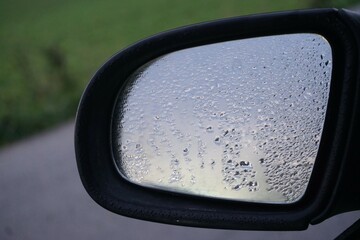 Nasser Autorückspiegel mit Spiegelung von grauem diesigem Himmel und Wassertropfenmuster bei Regen am Morgen im Herbst