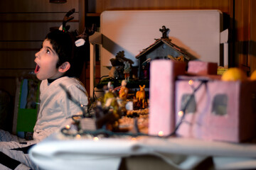 A child with a surprised and excited face, smiling and happy next to the Christmas Nativity Scene. He is wearing a reindeer headband and looking up. Selective focus. Scene full of great illusion