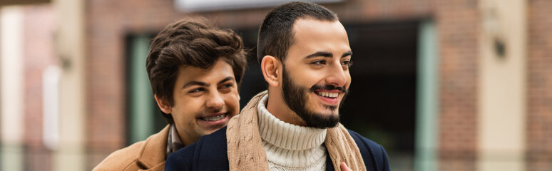 young and happy gay man smiling near bearded boyfriend outdoors, banner.