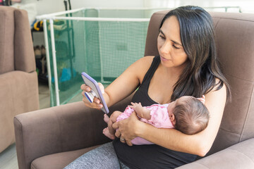 Mature woman with her newborn baby in her arms working at home from her smart phone