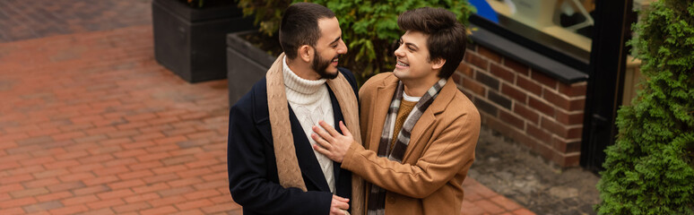 trendy gay man in beige coat touching chest of bearded boyfriend while smiling at each other outdoors, banner.