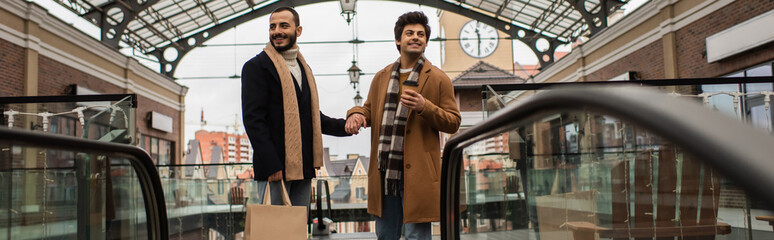 joyful and stylish gay men with shopping bags and coffee to go holding hands on city street, banner.