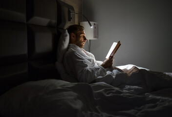 Midle-aged caucasian man relaxing in bed reading bestseller novel paper book with bedside lamp...