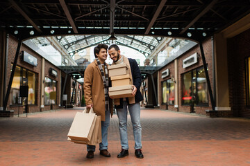full length of trendy gay couple with shopping bags and shoeboxes near building with stores on...