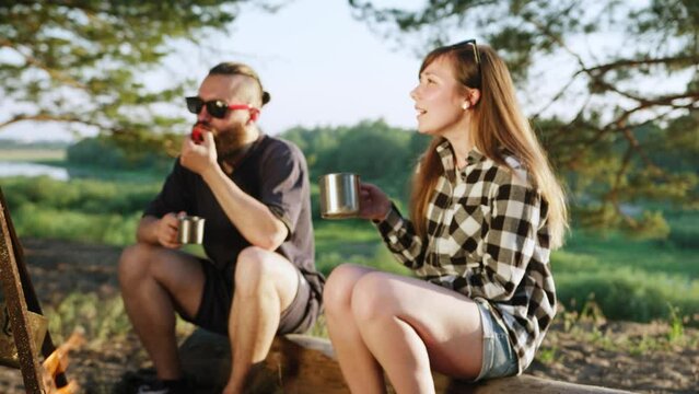 Young girl and hipster man eating tomato are sitting on log by the bonfire with friends and talking while drinking drinks