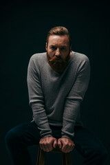Fototapeta premium Portrait of bearded man posing while sitting on chair isolated on black with shadow.