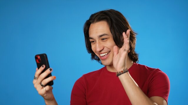 Happy Smiling Hispanic Latino Gender Fluid Young Man 20s In Red Shirt Talking Waving In Video Chat Call Using Mobile Cell Phone Isolated On Solid Blue Background Studio Portrait