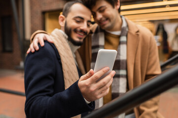 smiling gay man embracing bearded boyfriend using mobile phone outdoors on blurred background.