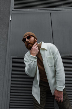 stylish man in beanie hat and sunglasses standing in white shirt jacket and scratching beard outside.