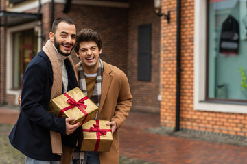 excited and stylish gay couple with Christmas presents laughing at camera near blurred building.