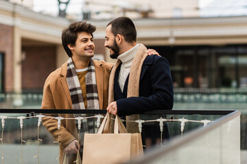 happy gays in trendy coats and scarfs holding shopping bags and looking at each other on urban street.