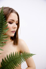 A young brunette girl of European appearance topless stands covering her chest with fern leaves on...