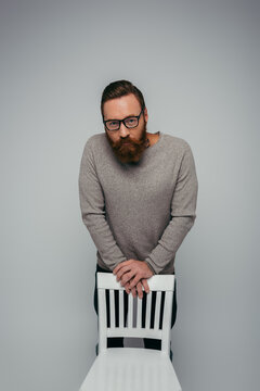 Bearded man in jumper looking at camera near white chair isolated on grey.