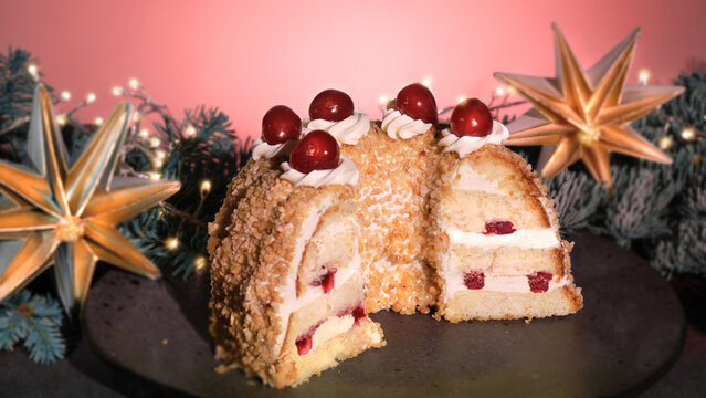German cake Frankfurter Kranz or Frankfurt Crown Cake. Bisquit with butter cream, cherry. Half of the cake on black plate. Black and pink background.Xmas fir twigs, paper star, garland with lights.