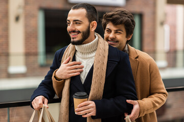 bearded gay in coat and scarf standing with takeaway drink near boyfriend embracing him outdoors.