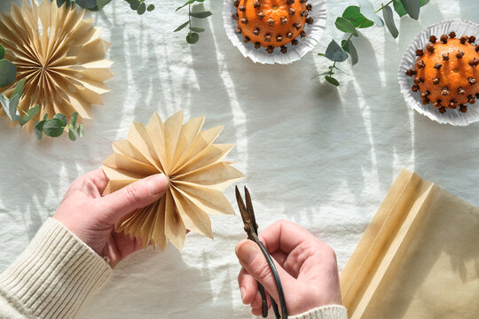 Fragrant pomander balls handmade from tangerines and cloves. Flat lay on off white textile tablecloth with aromatic wintertime green eucalyptus twigs. Hands making paper stars from brown baking paper.