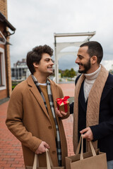 young gay man with shopping bags and Christmas present looking at happy bearded boyfriend.