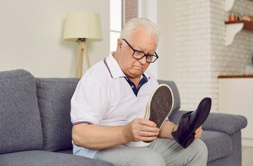 Senior man chooses which shoes to wear. Old man with dementia can't remember which shoes to put on. Chubby retired man sitting on sofa at home looking at classic and casual shoe he is holding in hands