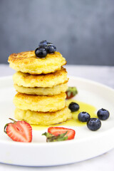 Cottage cheese pancakes with fresh strawberries, blueberries and honey on a white plate. Tasty breakfast food. Homemade syrniki. Coffee time. Grey background