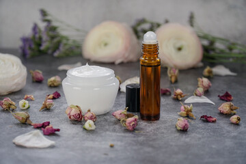  Natural under eye roll on in amber glass bottle and face cream in white packaging on grey background with flowers. Rose skin care. Cosmetics containers.             