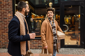 smiling gay man with purchases looking at boyfriend with paper cup near shop on city street.
