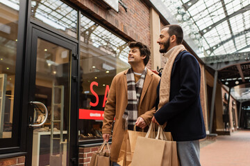 smiling gay couple with shopping bags looking at showcase on urban street.