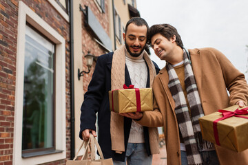 Obraz premium young gay man holding gift boxes near happy and trendy boyfriend with shopping bags on urban street.