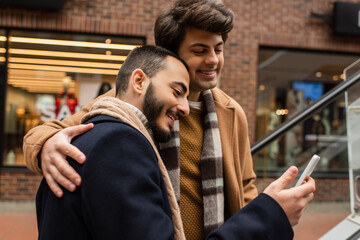 young and happy gay man in trendy outfit hugging boyfriend with smartphone on urban street.
