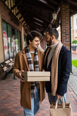 young gay man in checkered scarf holding shoebox near smiling boyfriend with shopping bags.