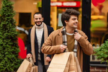 trendy gay man with shopping bags smiling and looking away near boyfriend and showcase on blurred...