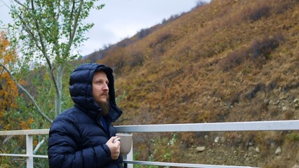 Fototapeta premium Warmly dressed tourist stands against the backdrop of high hill near the grill and drinks tea. Caucasian man in black jacket went outside to breathe fresh and cool autumn air and drink tea in nature.
