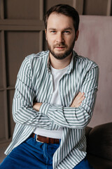 a young man with dark hair and a beard in a white t-shirt, a shirt with green stripes crossed his arms over his chest, close-up portrait