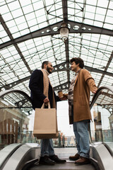 full length of smiling gay men with shopping bags and paper cup standing on escalator under transparent roof.