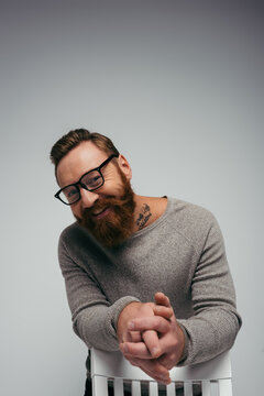 Positive bearded man in eyeglasses looking at camera near chair isolated on grey.