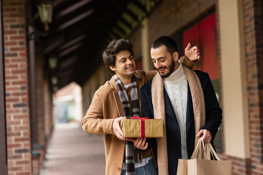 cheerful gay man presenting gift box to pleased and trendy boyfriend holding shopping bags on city street.