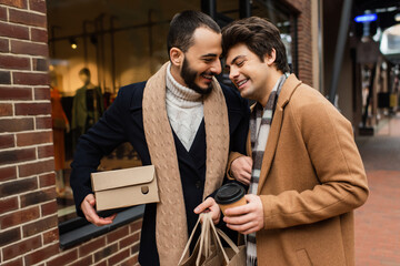 happy gay couple with purchases and paper cup standing near blurred showcase on city street.