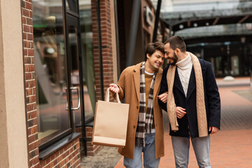happy gay man holding shopping bags while walking with bearded boyfriend on city street.