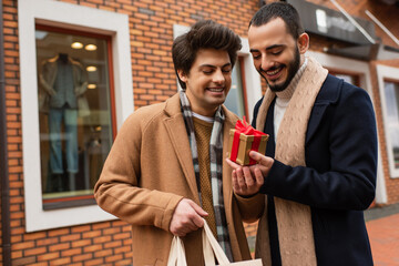 young gay man with shopping bag holding gift box near happy bearded boyfriend and shop on blurred background.