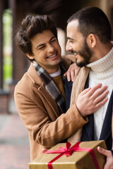 young and cheerful gay man touching chest of bearded boyfriend holding Christmas present outdoors.