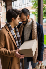 young gay man with closed eyes holding shoebox while standing face to face with happy bearded...