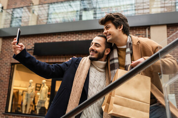 cheerful gay couple in trendy coats and scarfs taking selfie on smartphone on escalator.