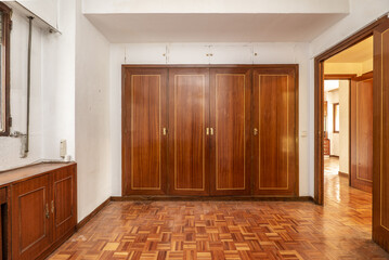 Empty room with a built-in wardrobe with four mahogany-colored wooden doors, a sideboard under the window and parquet floors of the same tone