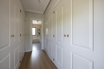 Empty room with white lacquered wooden built-in wardrobes forming a dressing room corridor with access to an en-suite bathroom
