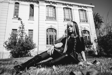 Portrait of a young and skinny model with long blonde and black hair, black clothes and black boots seated in the park in the day with old centuries building